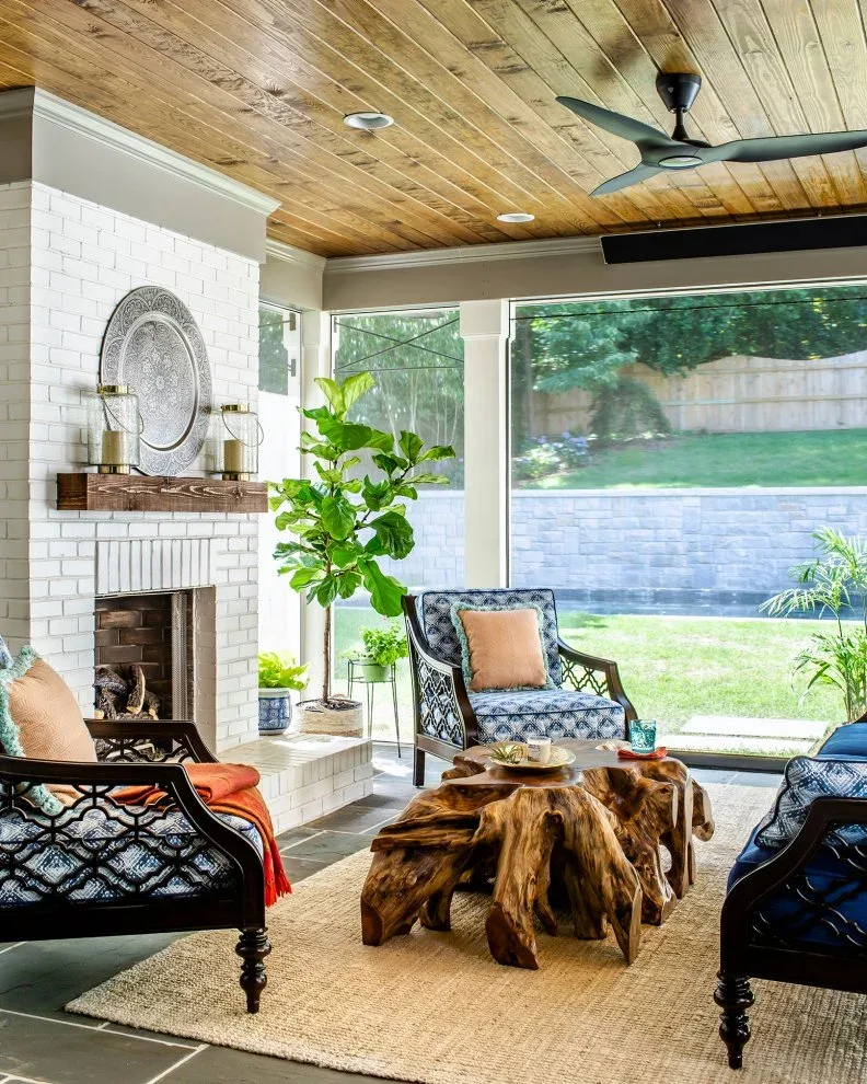 White brick fireplace in a glazed room with a wooden ceiling, patterned armchairs and irregular wooden table.