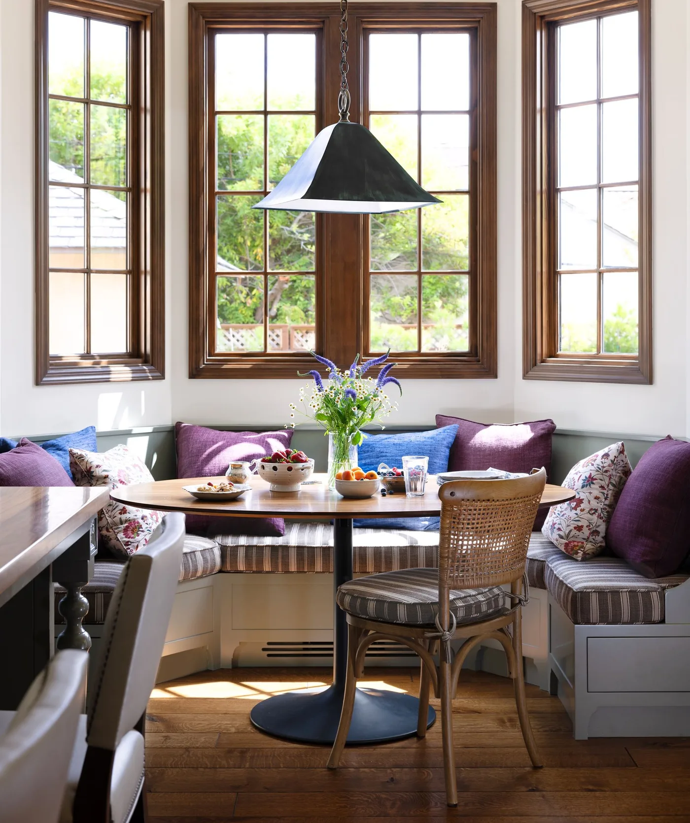Country dining nook with striped cushioned corner seating, purple and floral pillows, oval wooden table, woven-back chair and dark window trim.
