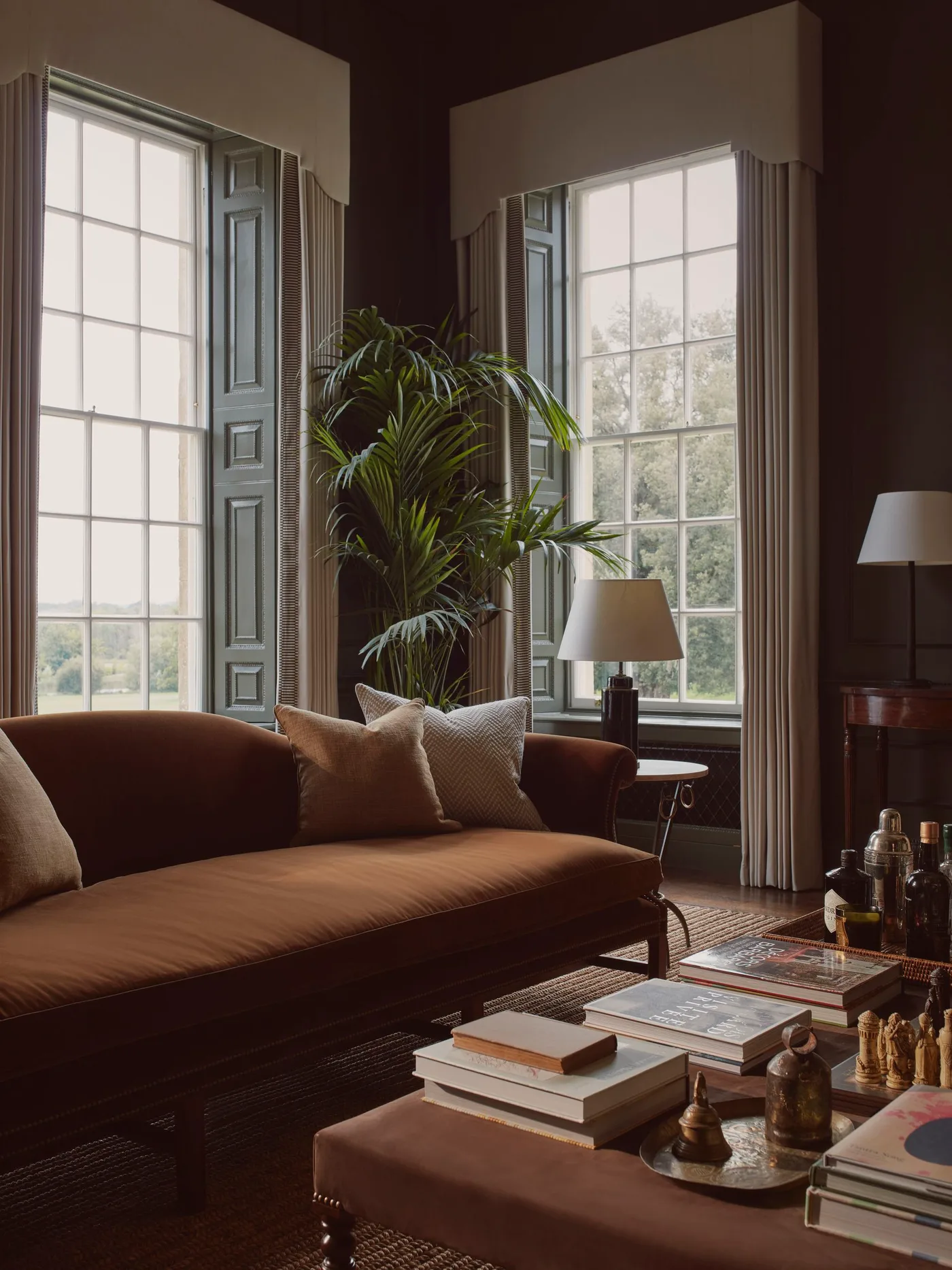 Deep brown living room with rust-toned sofa, tall shuttered windows, pale curtains and a leafy plant