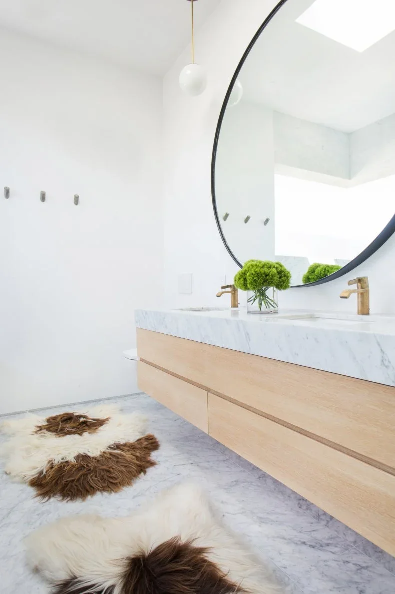 A large black round mirror hangs above a floating pale wood vanity with a veined white counter, gold-toned taps and small globe pendants