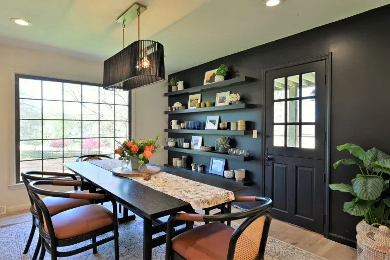 Dark floating shelves holding cups, plants and pictures beside a black dining table and orange seats