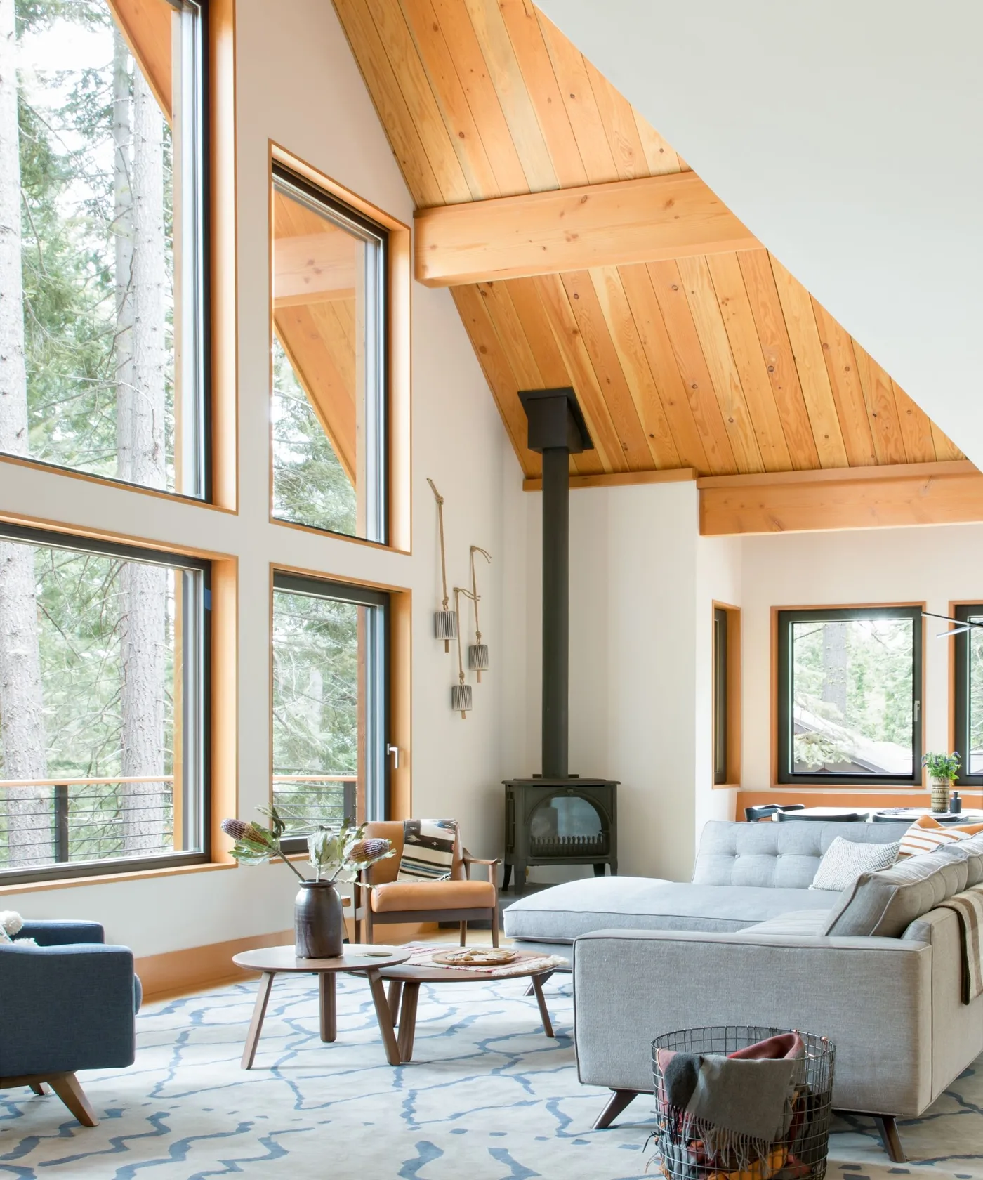 Black stove beside tall corner windows beneath a sloping wood ceiling, with pale seating nearby.