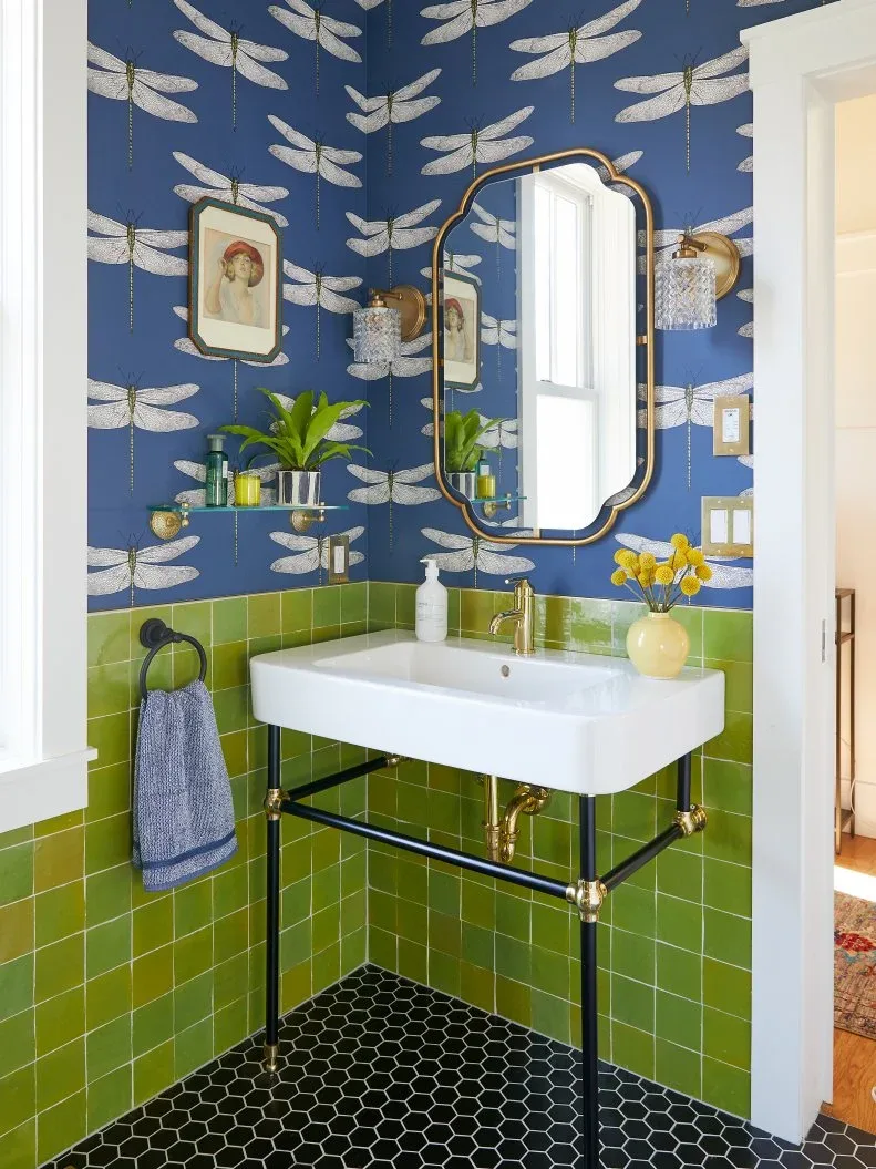 Powder room with blue dragonfly wallpaper, green wall tiles, white basin on dark console frame, shaped mirror, small plant and black hexagonal floor.