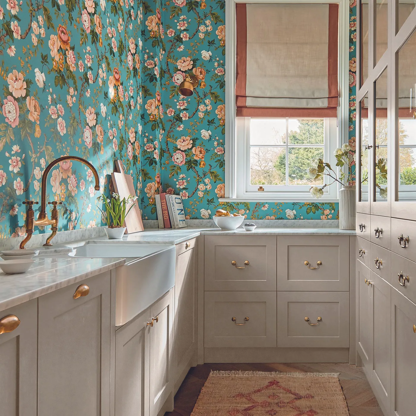 Pale framed kitchen with turquoise floral wallpaper, apron-front sink, gold-colored handles and a softly colored window blind.