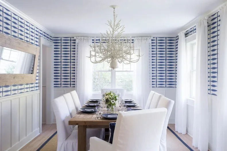 Dining room with blue and white patterned upper walls, pale lower panels, wooden table and white covered dining chairs.