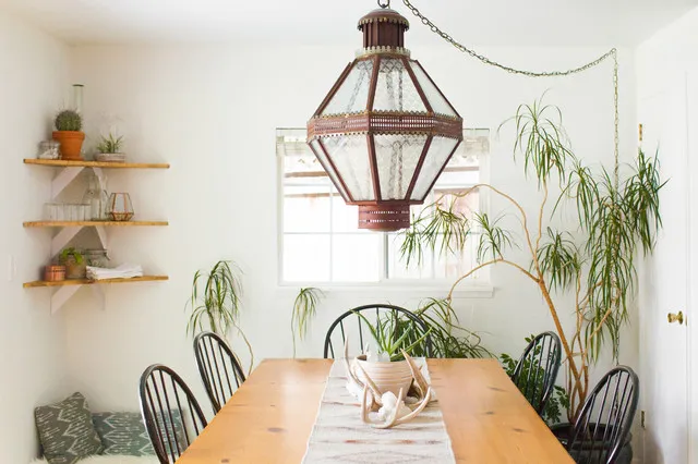 Timber dining table beneath a large framed lantern beside dark spindle chairs, shelves and a tall plant