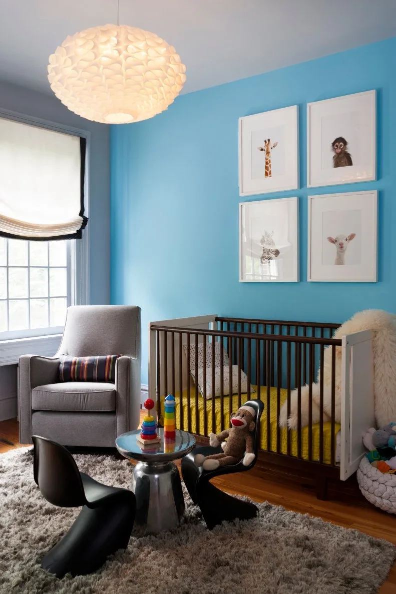 A wood nursery crib stands against a blue wall beside a pale armchair, framed animal pictures, a window shade and a rounded pale pendant