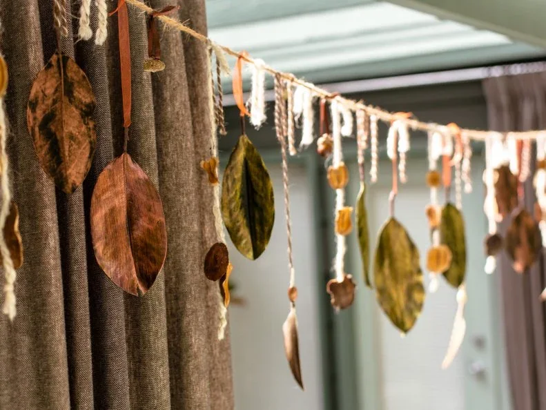Brown and green leaves hang at varied heights from a cord with pale and warm-colored ribbons in front of curtains