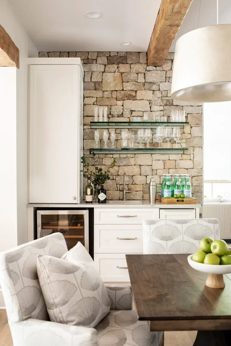 Two clear shelves holding stemware against a stone-textured wall above white cabinetry beside a wooden dining table