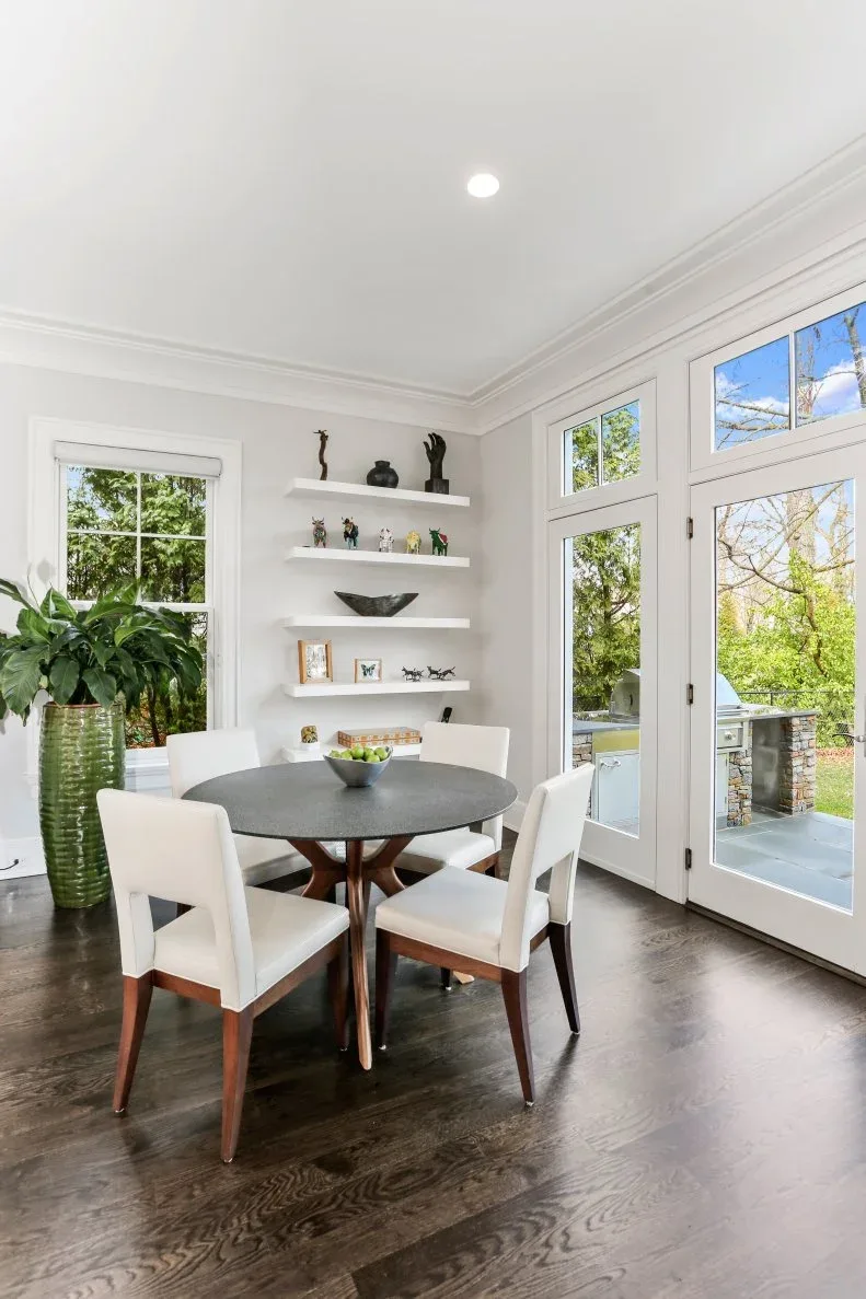 White floating shelves in a bright dining nook beside a dark round table, white chairs and glass doors