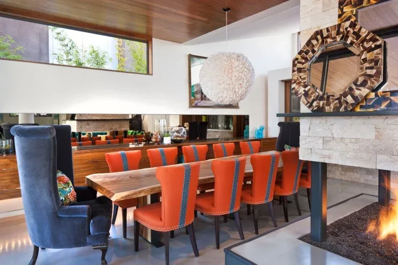 Orange dining chairs with blue vertical stripes beside a wooden table, a tall blue end chair, mirrored wall, and stone fireplace.