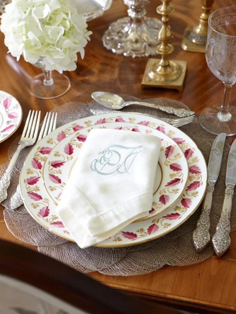 A pale monogrammed napkin rests across floral plates beside silver-colored cutlery, clear glasses and warm-toned candleholders