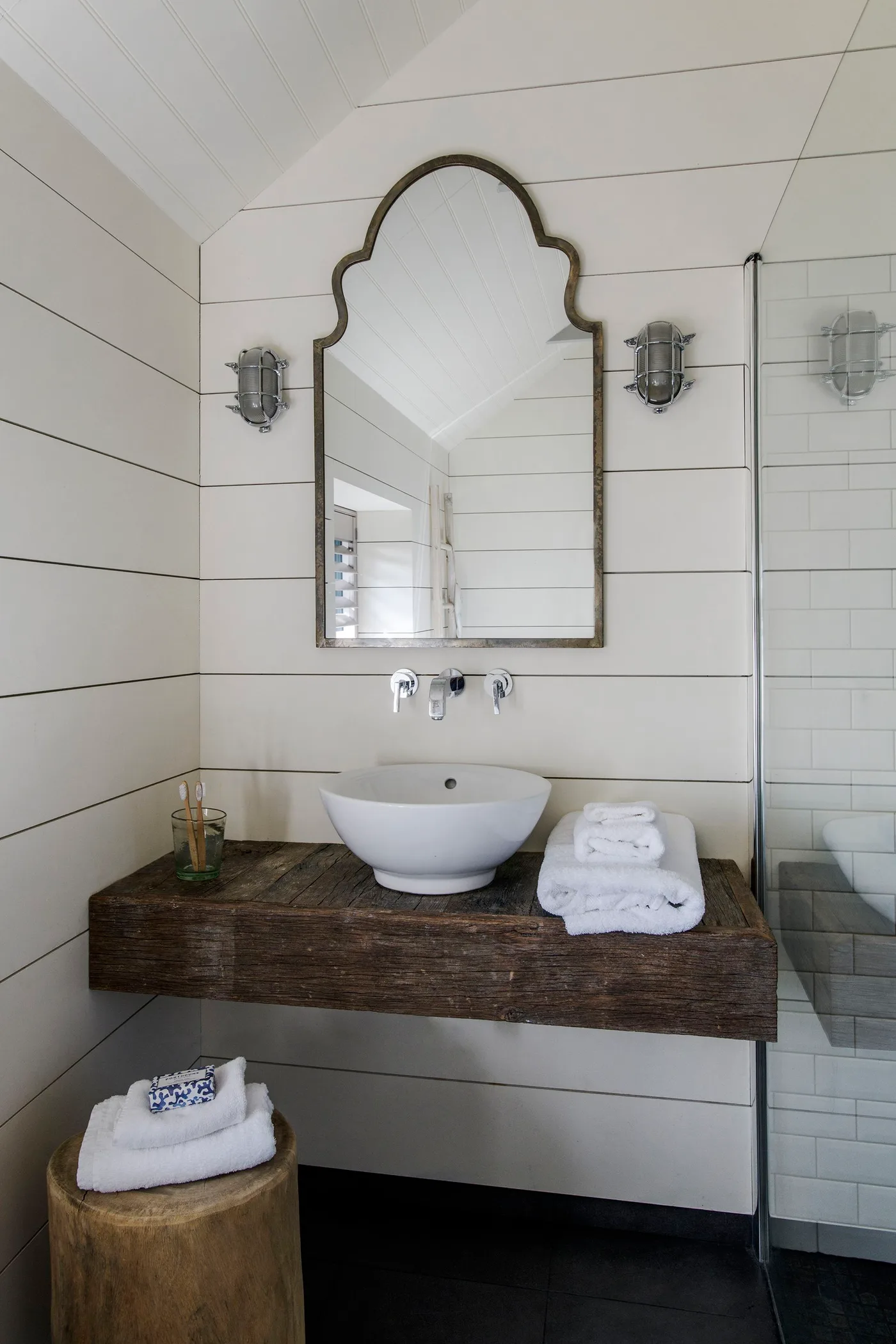 Country bathroom with pale horizontal wall boards, a white vessel basin on a thick wooden shelf and a tall shaped mirror beside a shower screen.