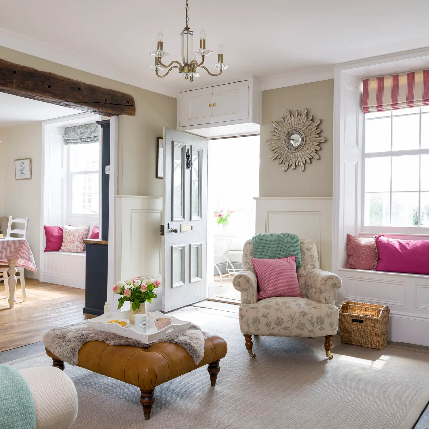 Country front door opening into a sitting room with pale paneling, a patterned armchair, window seat, baskets and a striped blind.