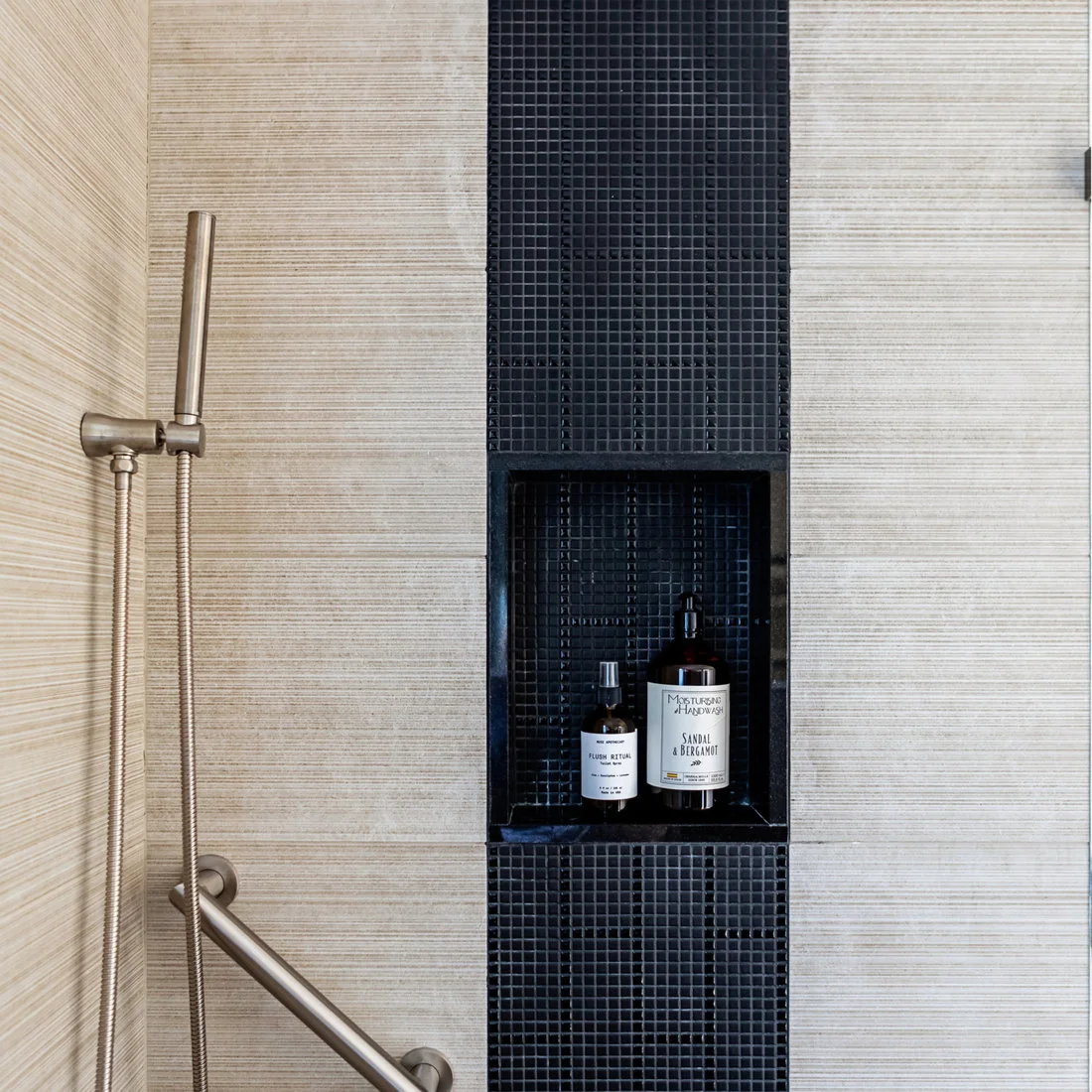 A dark small-tile shower strip surrounds a recessed bottle shelf between pale horizontally textured walls