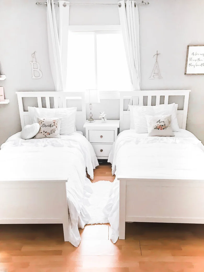 Two white single beds stand beside a shared white bedside chest below a window with white curtains and separate letter decorations above the headboards