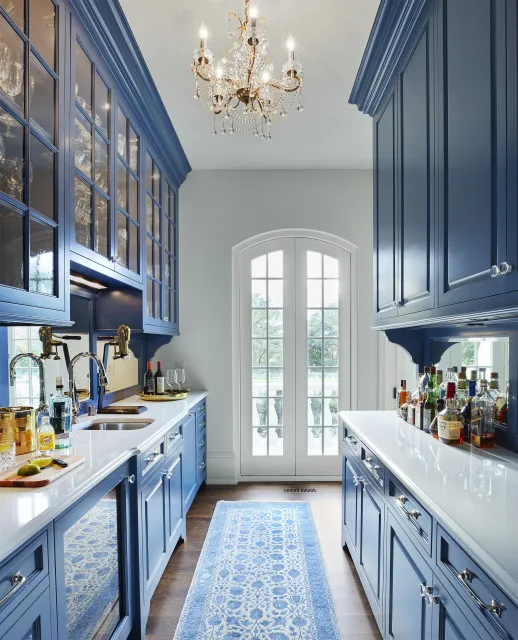 Blue cabinets run along both sides of a narrow kitchen with pale counters, glazed upper doors, a patterned runner and chandelier near white double doors
