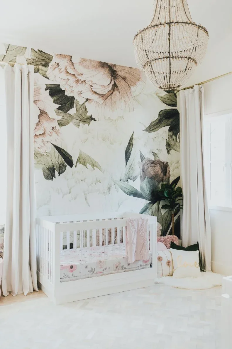 White crib against large pale floral wallpaper with darker leaves, lighter curtains, decorative pendant and staged pink fabrics.