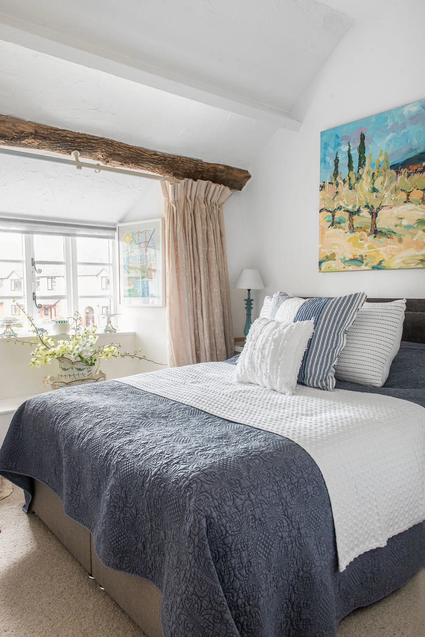 Bedroom with an exposed beam above a dormer window, pale walls, blue-gray bedcover, striped cushions and bright wall artwork.