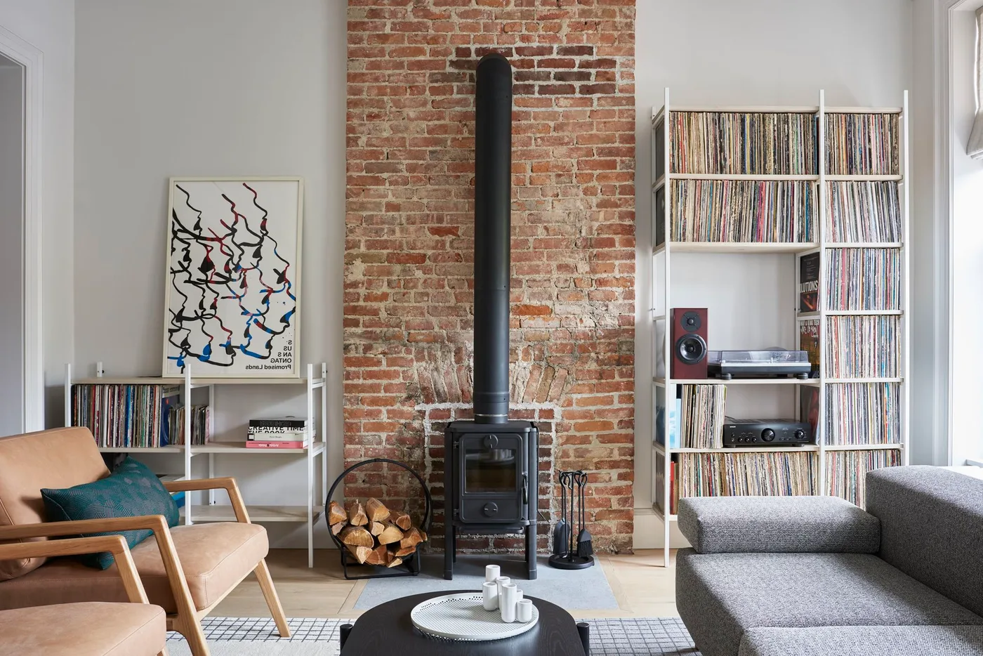 Brick chimney wall between record shelves in a living room with gray sofa, wood armchairs and black stove