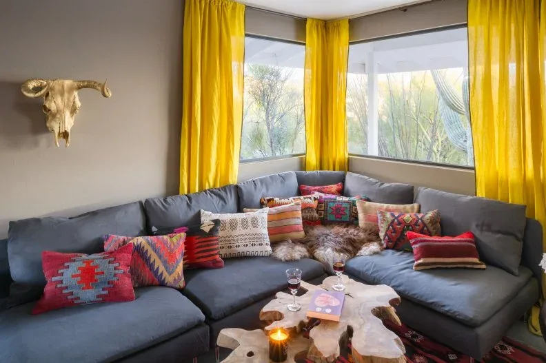 Gray sectional below yellow curtains with red patterned cushions and an irregular wooden coffee table.