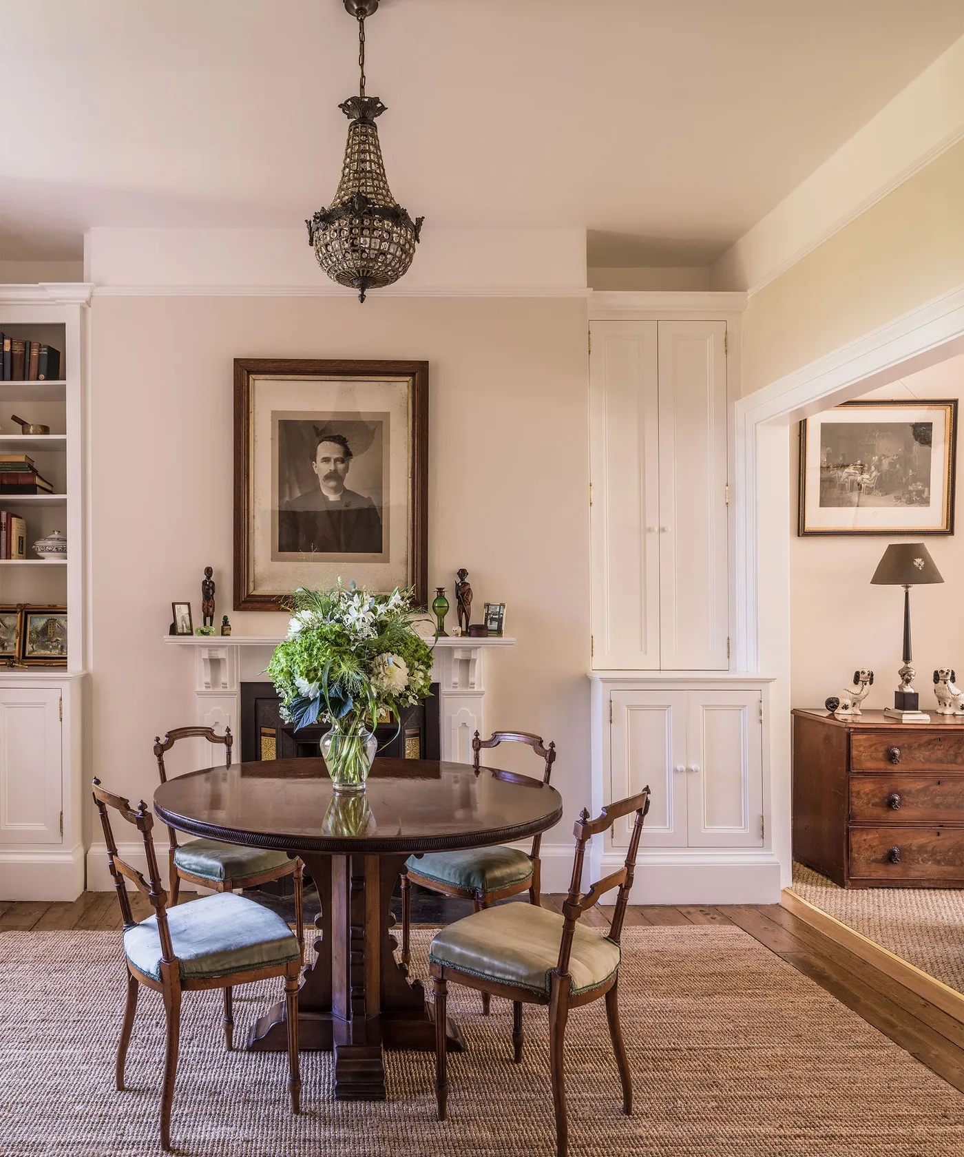 Cottage dining room with a round dark wooden table, curved wooden chairs, pale fitted cupboards, a fireplace and framed pictures.