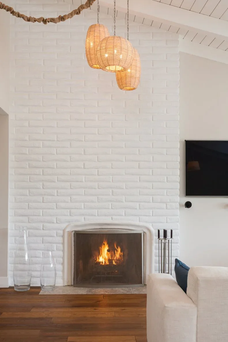 Tall white brick fireplace beneath a pitched paneled ceiling and three woven pendant shades.