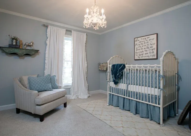 Soft blue nursery with white spindle crib, chandelier, pale armchair, curtains and wall shelf