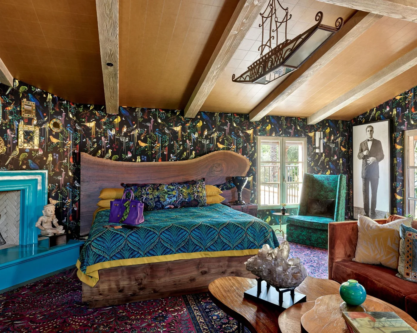 Dense botanical wallpaper surrounds a curved wooden headboard, patterned bedding, colorful chairs and a turquoise fireplace