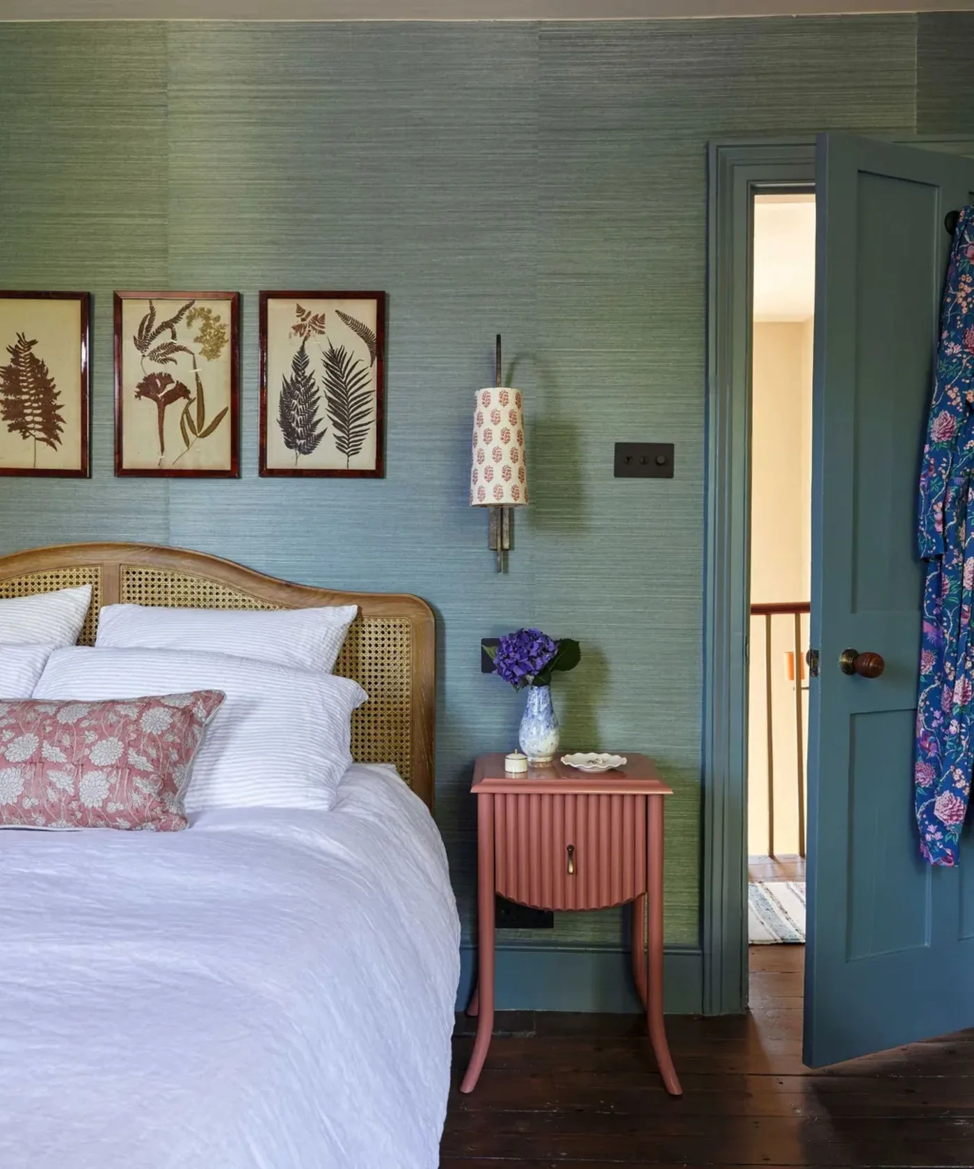 Blue textured bedroom wall with a woven curved headboard, white bedding, pink bedside cabinet and botanical pictures.