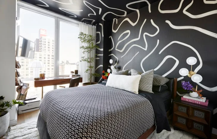 A bedroom has black walls and ceiling with broad white abstract lines above a bed with finely patterned bedding beside large windows and a wood desk