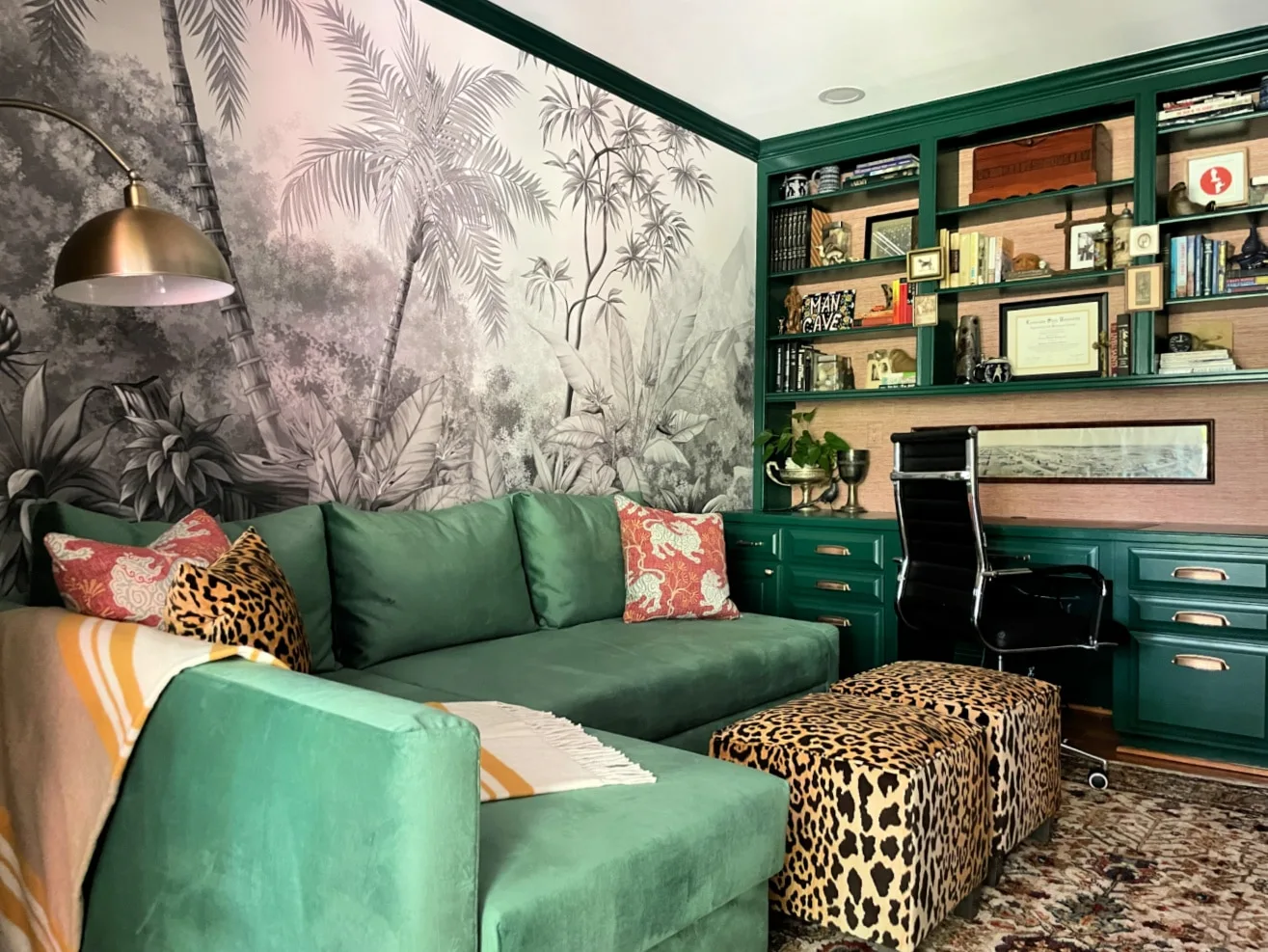 Green office shelves and drawers with warm metal pulls surround a desk beside green seating and botanical wallpaper