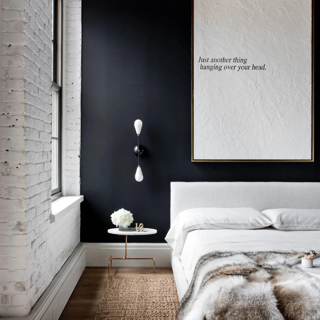 A pale upholstered bed sits beside a white painted brick wall and window against a dark wall with a slim black bedside light
