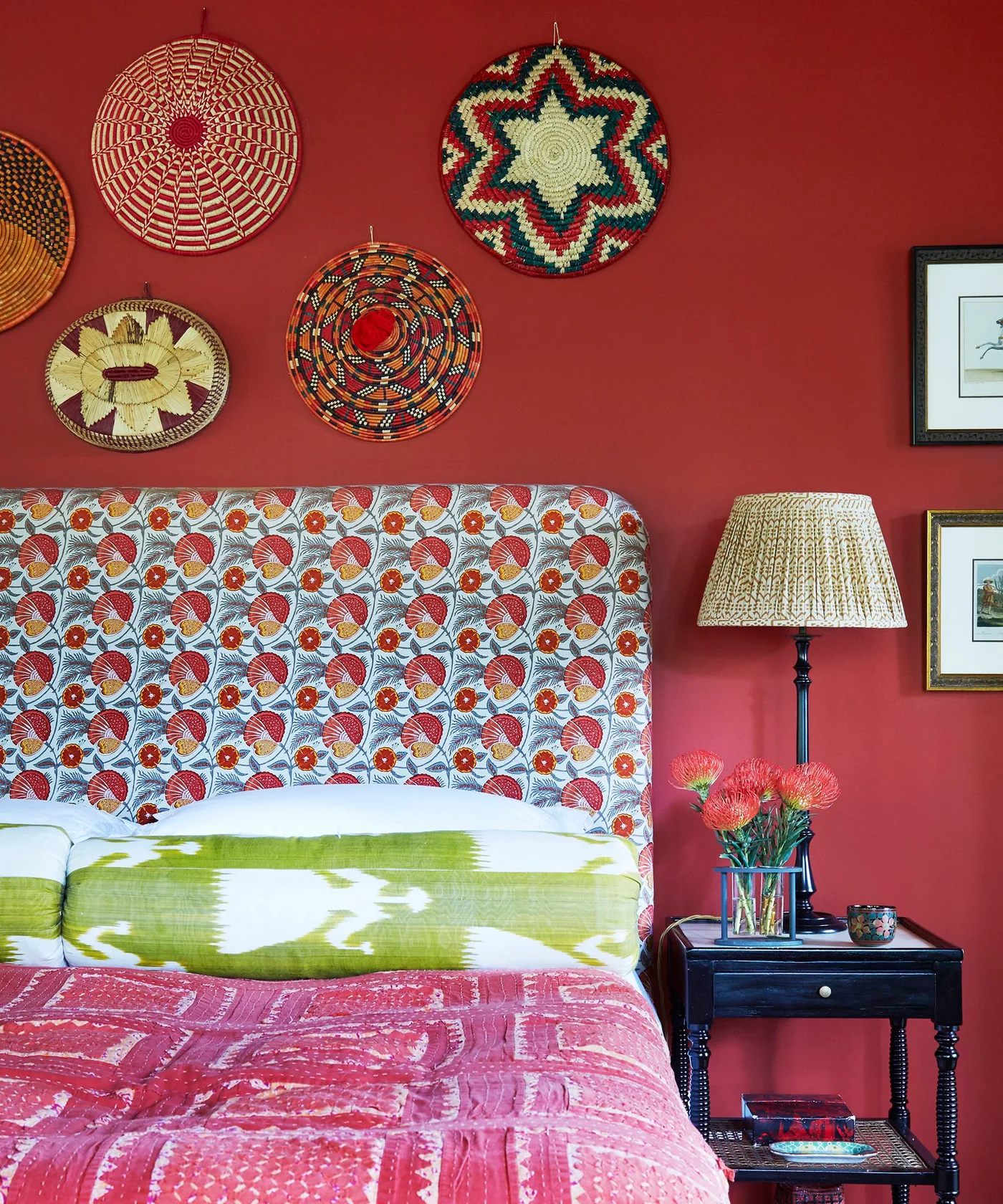 Bedroom with red wall, woven decorative plates, floral upholstered headboard, green and pink bedding and dark turned-leg bedside table.