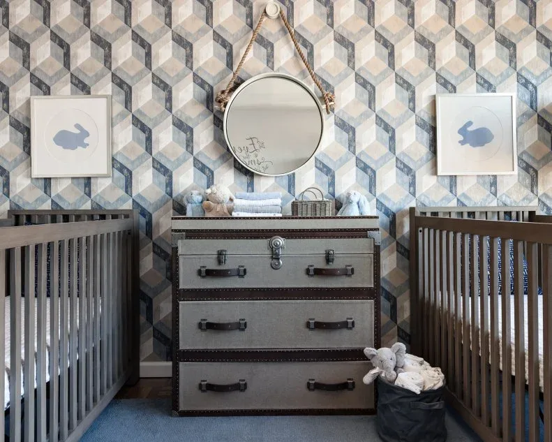 Two dark cribs flanking gray drawer unit against blue-gray geometric wallpaper with framed animal silhouettes and round mirror.