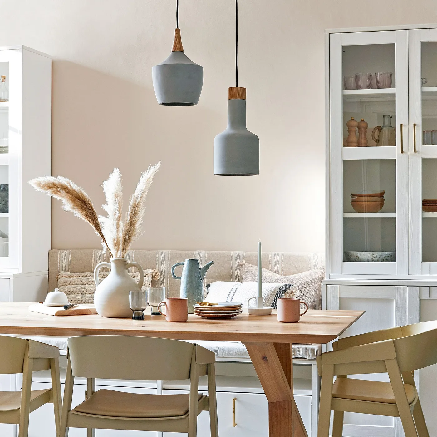 Pale dining room with a wooden table, cushioned bench between tall glazed cupboards, green-gray chairs and two blue-gray pendant lights.