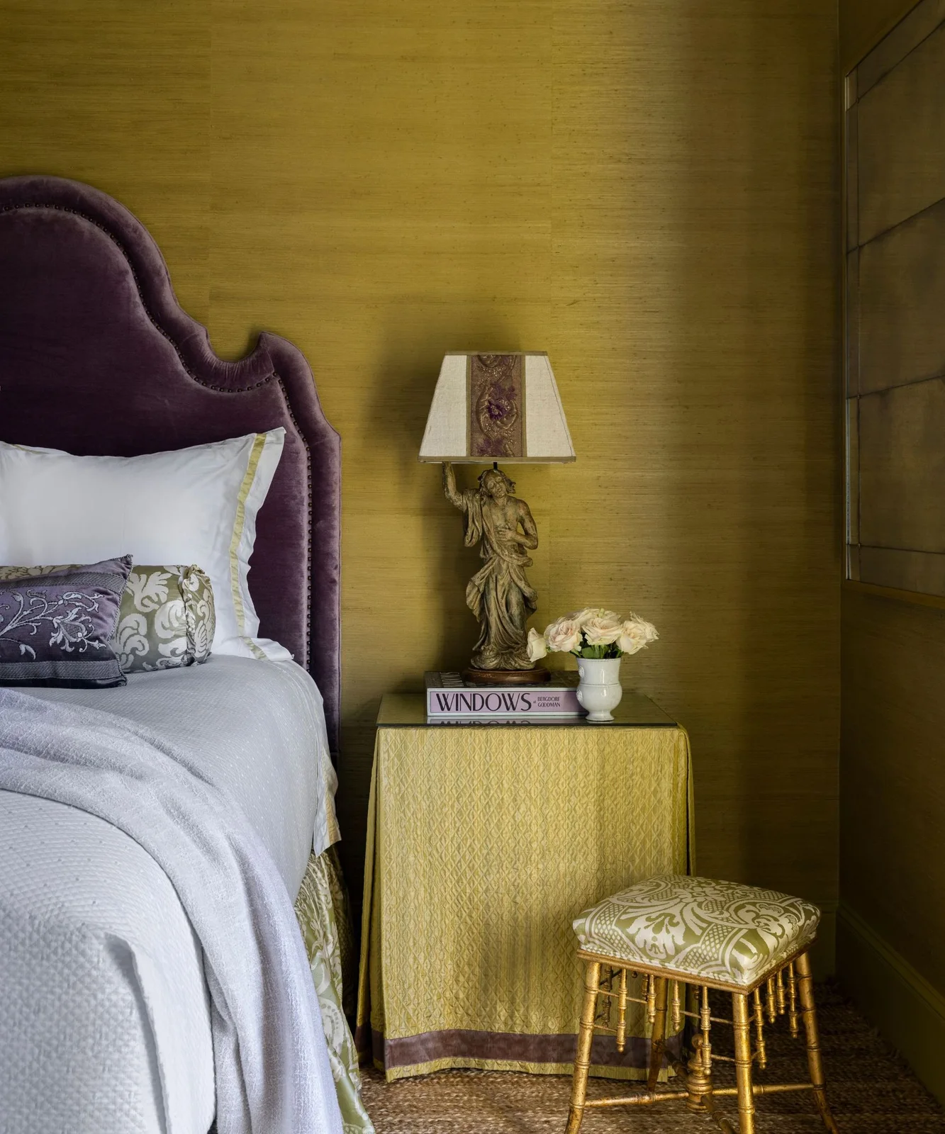 Yellow-green walls frame a tall curved purple headboard, patterned cushions, a decorative bedside lamp and a green floral stool cushion