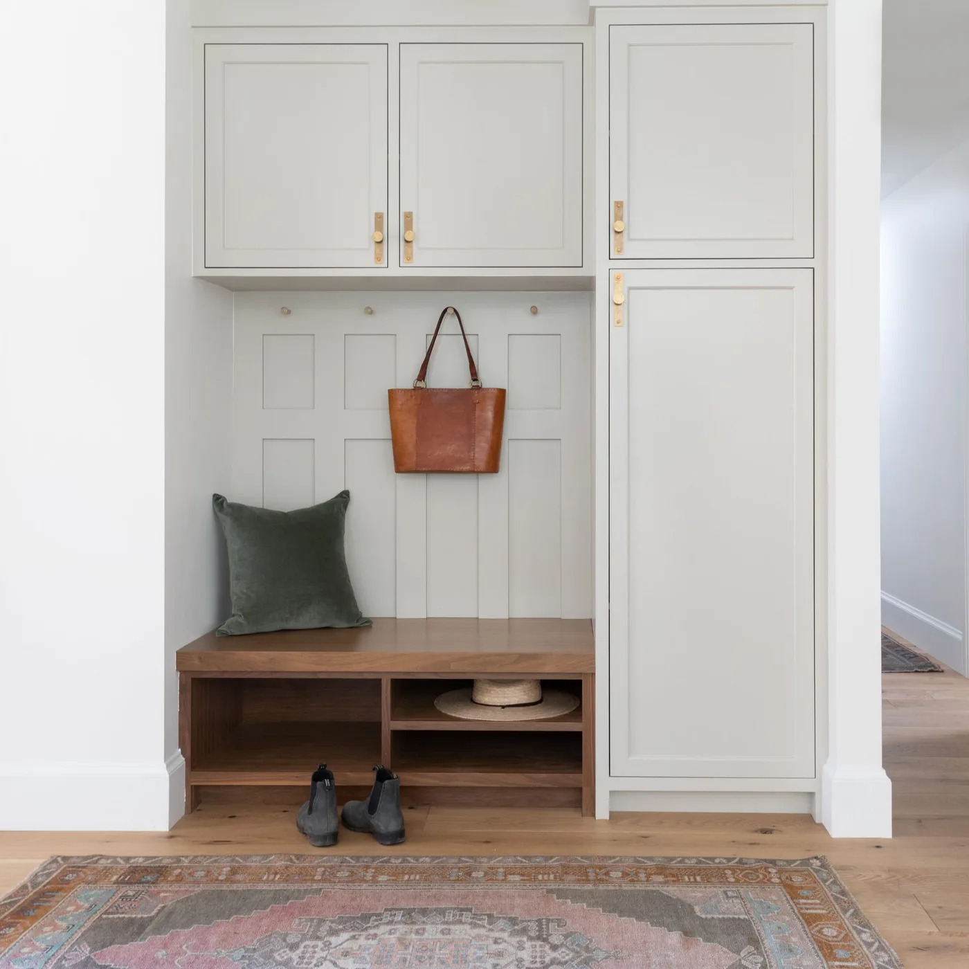 Pale recessed mudroom with wooden bench and open shoe shelves, upper cupboards, side tall cabinet and hanging bag.