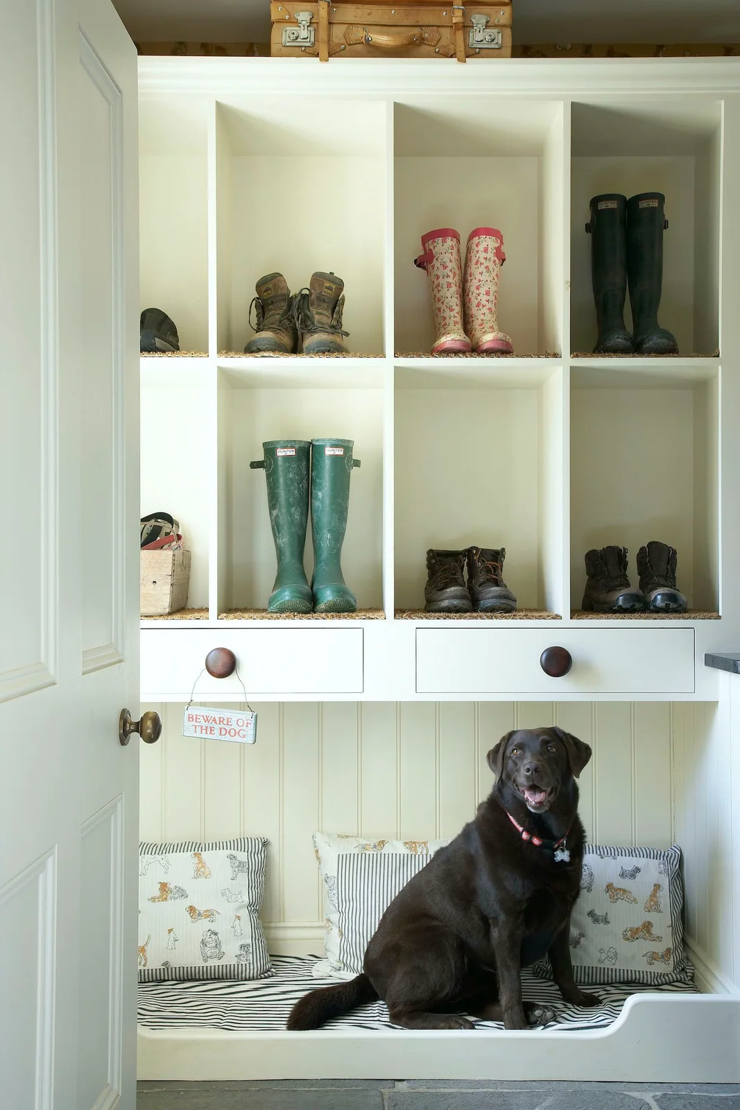 Pale boot-storage cubbies and two drawers sit above a lower cushioned nook beside a paneled door, with a dog seated on the cushion
