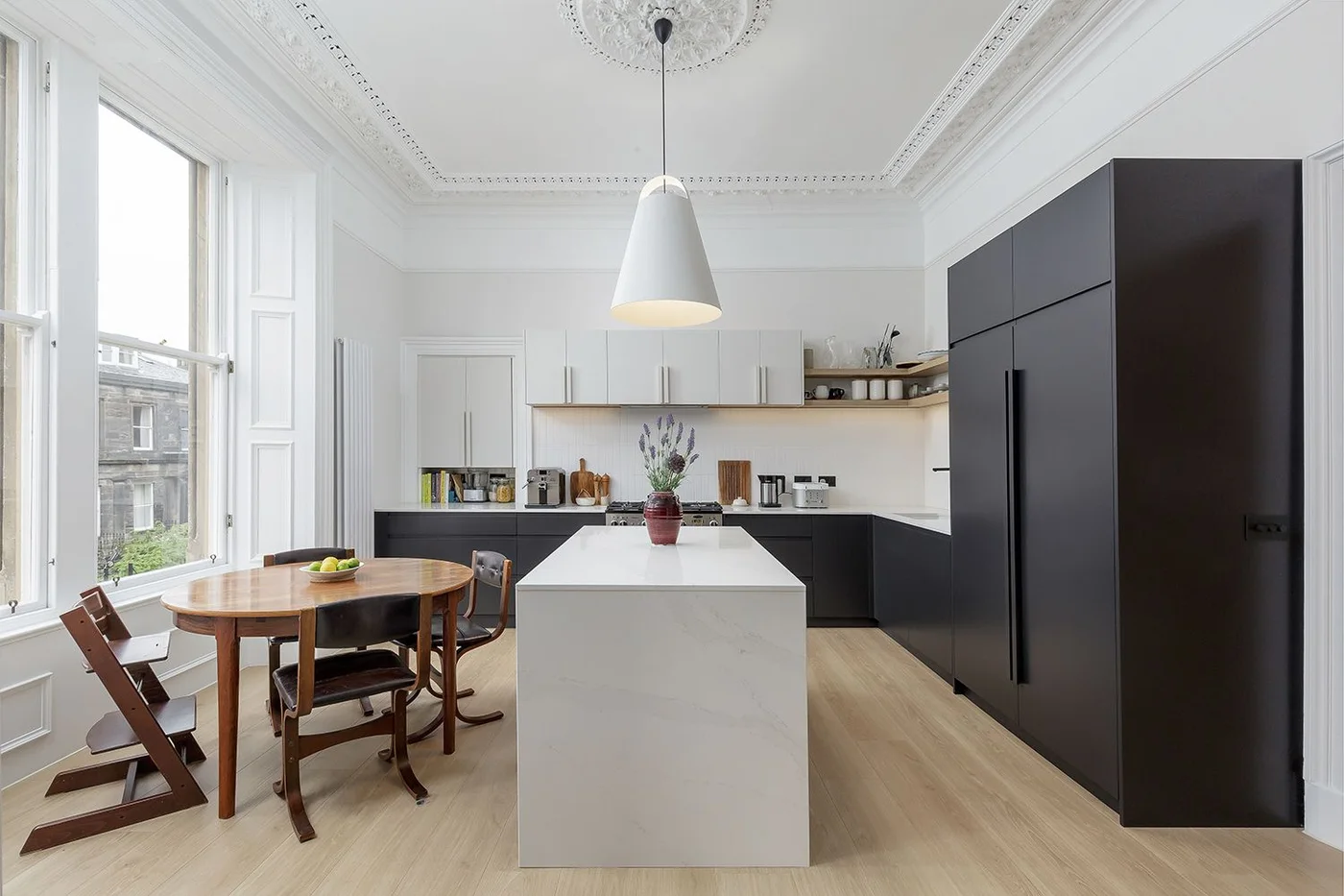 A narrow pale kitchen island stands beside dark tall cabinets and lower cupboards beneath white upper storage near a small wood dining table and broad window