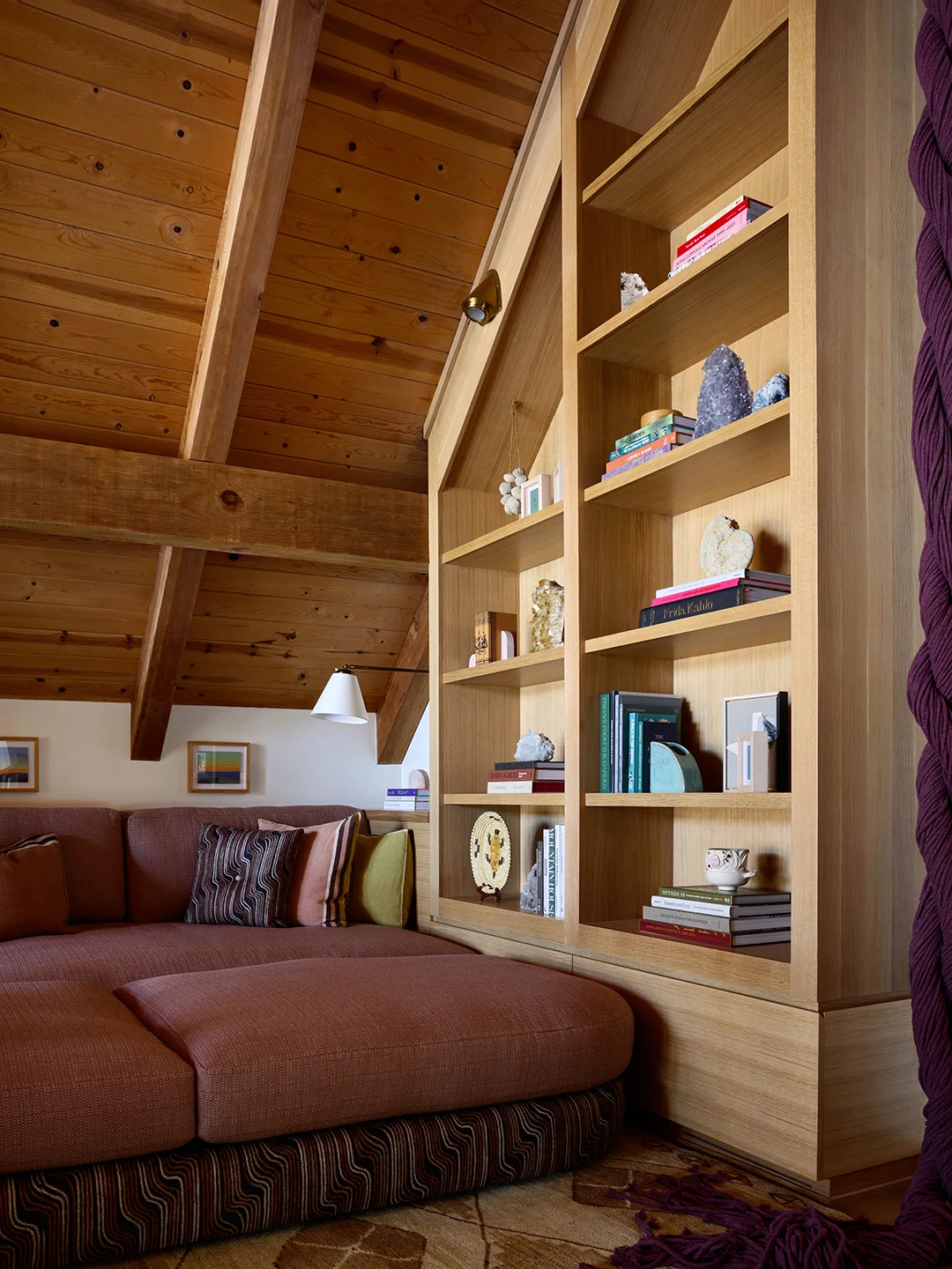 Wooden shelves follow a sloping roof beside a rust-colored sofa and ottoman under exposed wooden framing