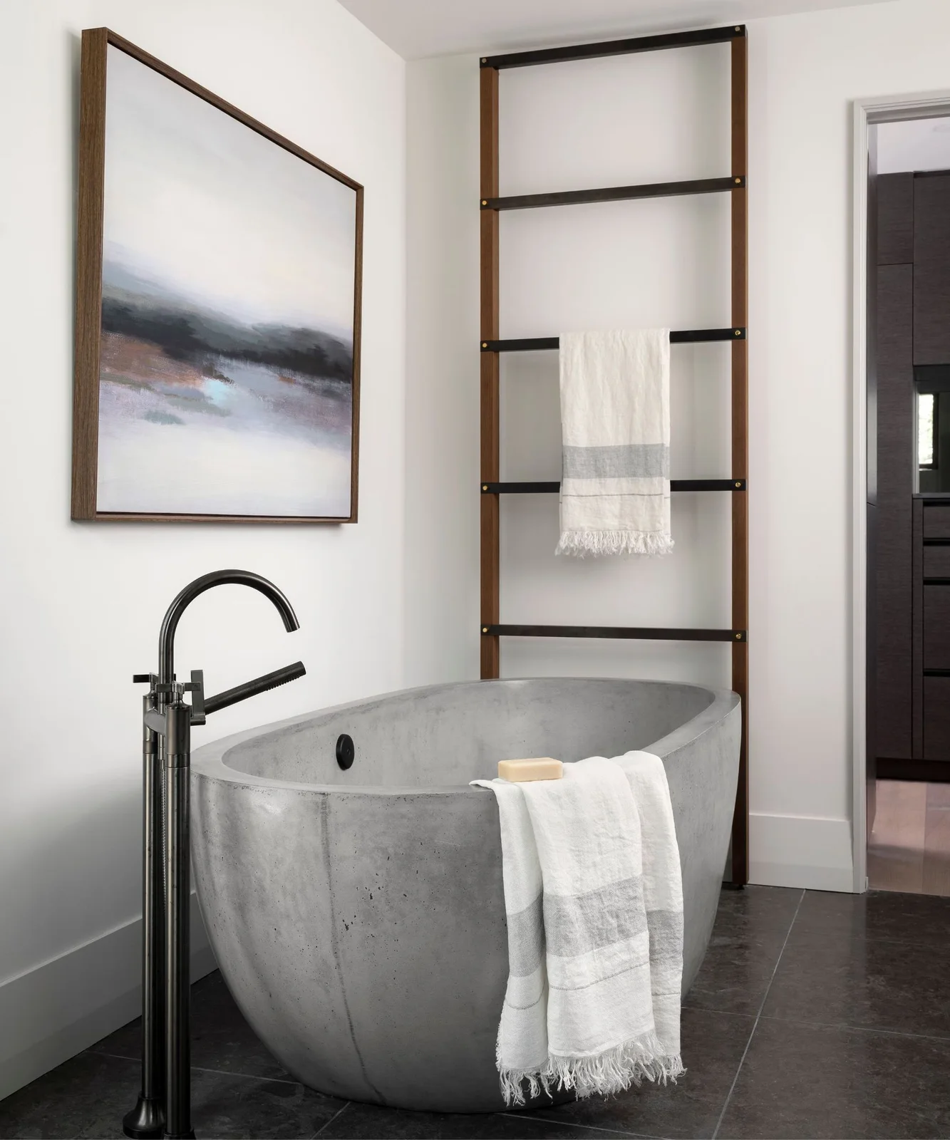 A gray freestanding tub sits beside a black floor-mounted faucet beneath framed artwork and a tall wood-and-black towel ladder