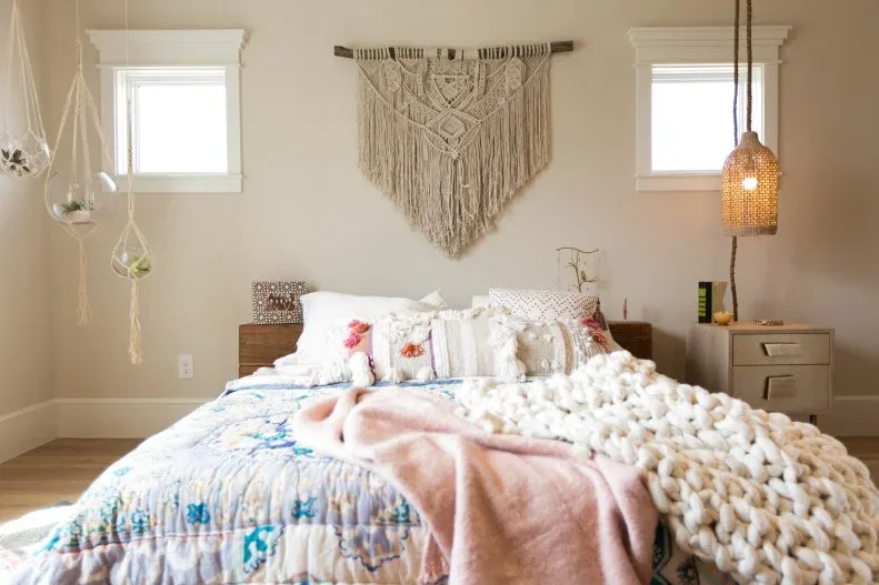 Bedroom with fringed woven wall hanging, small hanging planters beside the window, woven pendant shade and layered white, blue and pink bedding.