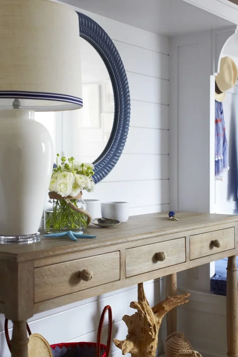 Coastal entry detail with wooden drawer console, round blue mirror, pale lamp, flowers and white paneled wall.