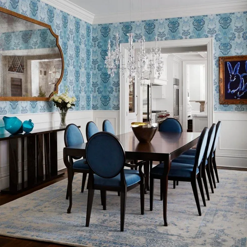 Blue oval-backed dining chairs around a dark table beside blue patterned wallpaper and a clear chandelier.
