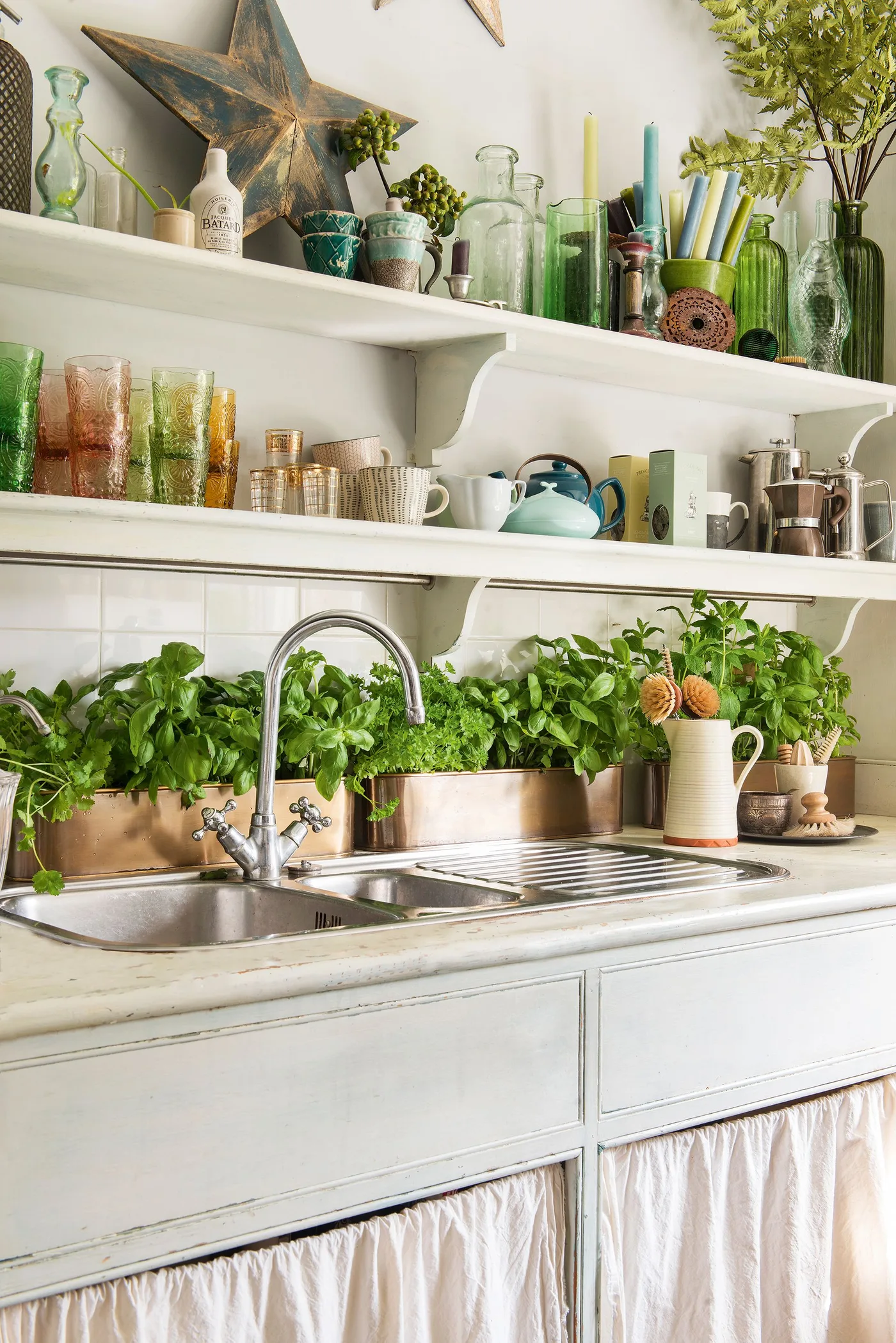 White kitchen shelves with green glass bottles, colorful tumblers, a star ornament and herbs near the sink.