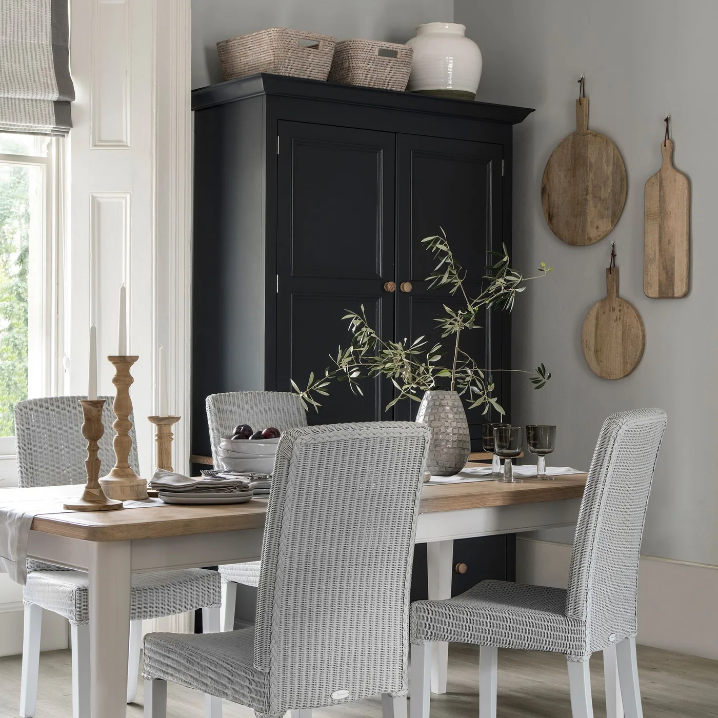 Cottage dining room with a tall black paneled cupboard, pale wooden table, light woven chairs and wooden cutting boards on the wall.