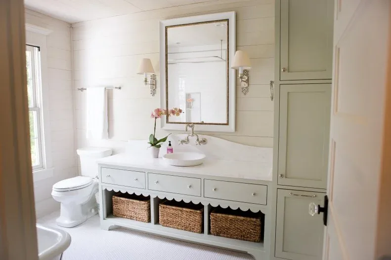 Soft green bathroom vanity with scalloped lower edge, three woven baskets, side cupboard, tall mirror and white horizontal paneling