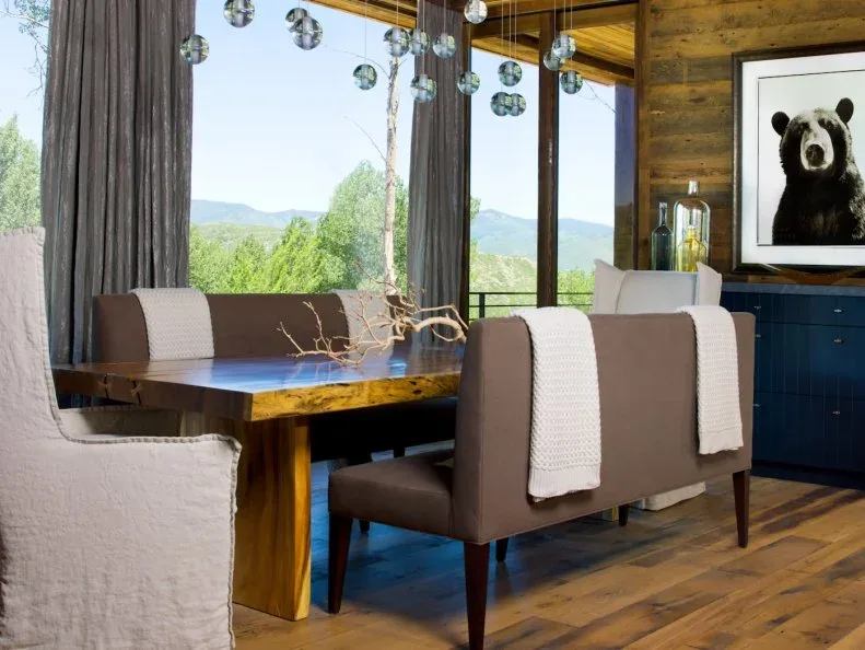 Dark blue fitted lower cabinets beneath wooden wall paneling and a framed bear picture, beside a thick wooden dining table, upholstered chairs, and tall windows.