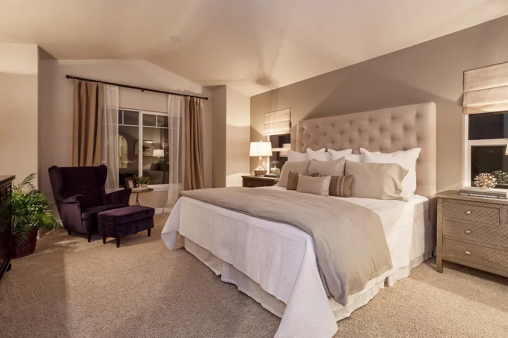 A tufted taupe-beige bed stands against a warm neutral wall above pale carpet